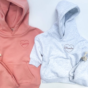 May include: Two hooded sweatshirts with embroidered hearts and names. One is pink with the name "Joanie" and the other is gray with the name "Phoebe".