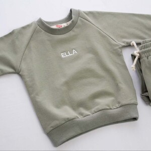 Pode incluir: Um conjunto de beb&ecirc; de duas pe&ccedil;as em verde-s&aacute;lvia com uma camiseta de manga comprida e cal&ccedil;as de moletom combinando. A camiseta tem um nome bordado em branco "ELLA" na frente.