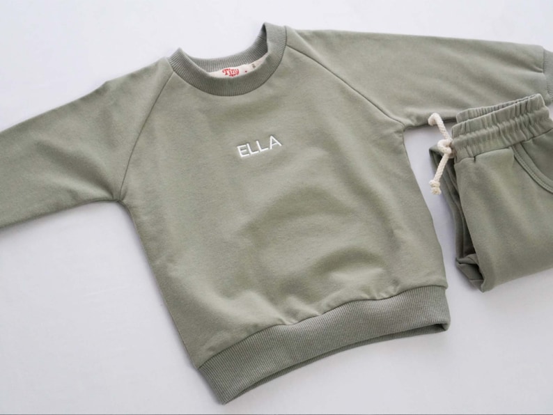 Pode incluir: Um conjunto de beb&ecirc; de duas pe&ccedil;as em verde-s&aacute;lvia com uma camiseta de manga comprida e cal&ccedil;as de moletom combinando. A camiseta tem um nome bordado em branco "ELLA" na frente.