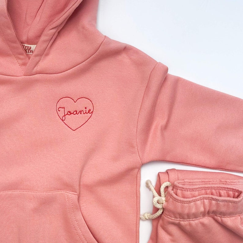 May include: A pink hoodie with a heart embroidered on the front that says "Joanie". The hoodie is paired with matching pink sweatpants.