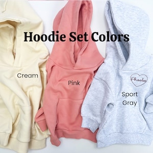 May include: Three hooded sweatshirts in cream, pink, and gray. The text "Hoodie Set Colors" is displayed above the sweatshirts. The cream sweatshirt has the word "Cream" written below it. The pink sweatshirt has the word "Pink" written below it. The gray sweatshirt has the words "Sport Gray" written below it and the word "Phoebe" written in a heart shape.