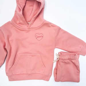May include: A pink hooded sweatshirt and matching sweatpants set with a red embroidered heart and the name "Joanie" inside.