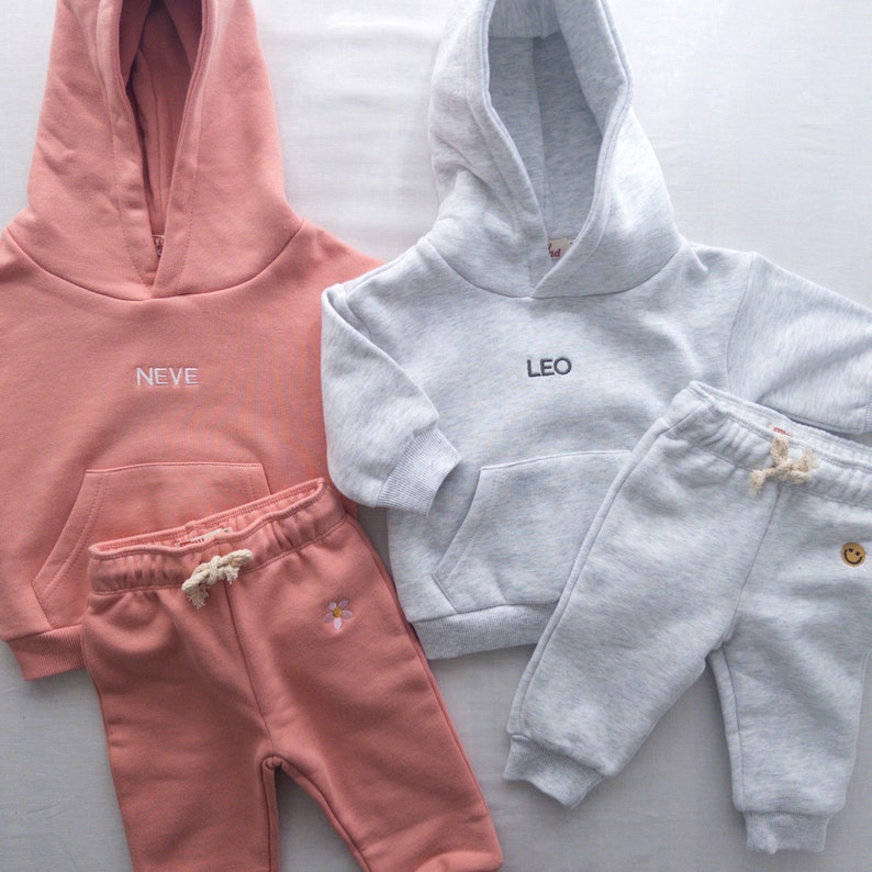 May include: Two sets of children's clothing. One set is pink with a hoodie that says "NEVE" and matching sweatpants with a flower embroidery. The other set is gray with a hoodie that says "LEO" and matching sweatpants with a smiley face embroidery.