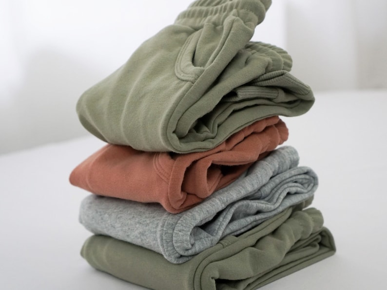 Pode incluir: Uma pilha de quatro pares de cal&ccedil;as de moletom em diferentes cores, incluindo verde, cinza e laranja.