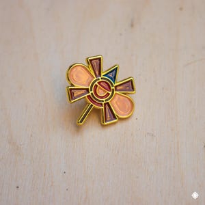 May include: A gold-toned pin with a geometric design featuring a central circle with concentric rings in red, orange, and blue. The design is surrounded by a stylized flower shape with pink and burgundy petals.