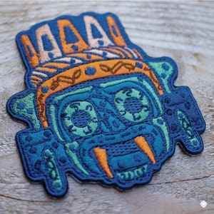 May include: Embroidered patch featuring a blue and green stylized face with orange teeth and a crown. The face has a geometric design with intricate details.