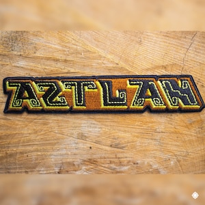May include: A brown and yellow embroidered patch with the word "AZTLAN" in black lettering.