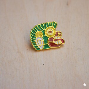 May include: A gold-colored enamel pin with a green, red, white, and yellow design. The design is a stylized face with a large mouth and a single eye.