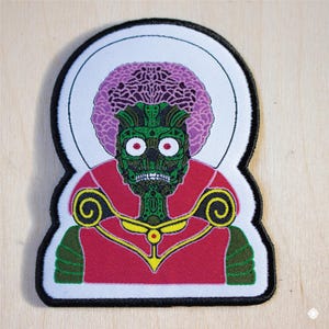 May include: A white, black, and green embroidered patch featuring a cartoon alien with a purple afro, red eyes, and a green face. The alien is wearing a red and gold robe with a yellow collar.