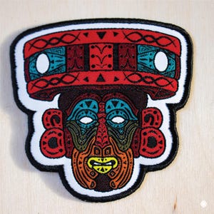 May include: A colorful embroidered patch featuring a stylized face with intricate details. The face is surrounded by a red, blue, and white patterned border.