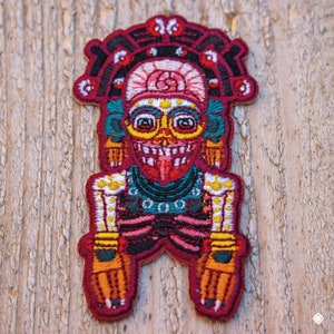 May include: Embroidered patch featuring a colorful, detailed depiction of a deity with a toothy grin, wearing a crown and ornate clothing. The patch is set against a wooden background.