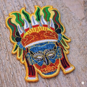 May include: A colorful embroidered patch depicting a stylized face with a headdress. The face is blue with yellow, orange, and red details. The headdress is green, yellow, and white.