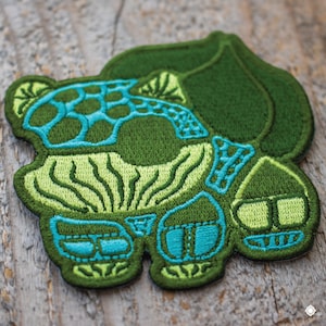 May include: Green and blue embroidered patch of a stylized mushroom with a leaf on top.
