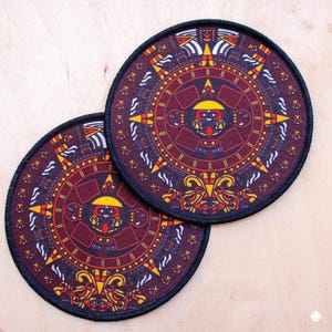 May include: Two round patches with a detailed Aztec calendar design. The patches feature a burgundy background with yellow, orange, and white accents. A black border surrounds the circular design. The patches are likely for clothing or accessories.