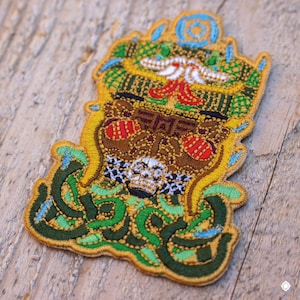 May include: Embroidered patch with a detailed design featuring a skull, intricate patterns, and a gold border. The patch incorporates green, brown, red, white, and blue colors, creating a vibrant and ornate aesthetic. The patch is likely for clothing or accessories.