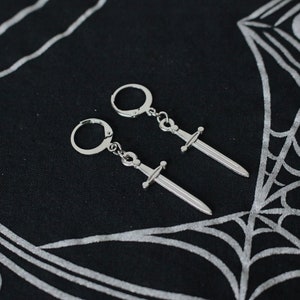 May include: A pair of silver sword earrings. The earrings are hanging from a black background with a silver design.