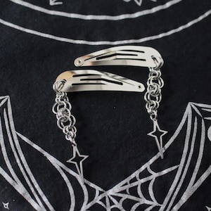May include: Two silver hair clips with chain and star charms. The clips are on a black background with a white web and crescent moon design. The hair accessories are a stylish addition to any hairstyle.