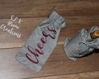 May include: Two gray wine bottle bags. One bag is upright with the word "Cheers" in burgundy script. The other bag is wrapped around a wine bottle. The bags are made of a textured fabric. The text "SLK Home Creations" is in the upper left corner.