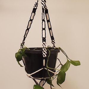 May include: A black plastic plant pot held by a macrame plant hanger made of black and white cord. The hanger features a metal ring at the top for suspension and a knotted design that secures the pot, with a green trailing plant inside the container.