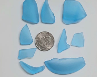 May include: A collection of ten sea glass pieces in various shapes and sizes. The sea glass is a light blue color and has a frosted finish. The sea glass is arranged on a white surface.