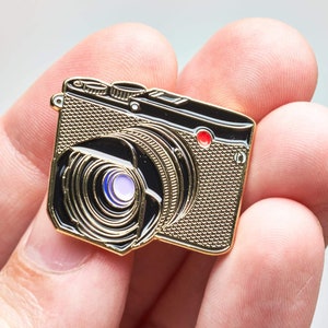 Puede incluir: Un pin de cámara dorado con detalles en negro y rojo. El pin presenta un diseño detallado de una cámara vintage, que incluye la lente, los botones y el cuerpo. La lente tiene un centro morado y azul.