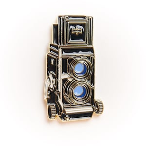 Puede incluir: Un pin de esmalte negro y dorado con la forma de una cámara réflex de doble objetivo vintage. El pin presenta dos círculos azules que representan las lentes y detalles intrincados. Un accesorio elegante para los entusiastas de la fotografía.