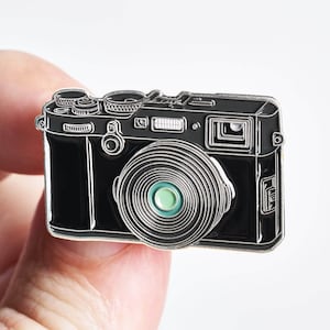 Könnte beinhalten: Schwarzer und silberner Kamera-Emaille-Pin mit grünem Objektiv.