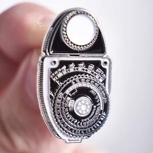 May include: A silver-toned metal pin shaped like a vintage light meter. The pin has a black face with a circular dial and a white circle in the center. The dial has numbers and markings around the edge.