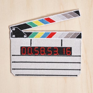 Puede incluir: Parche bordado de claqueta de cine con base blanca, detalles negros y sección superior colorida. La claqueta muestra el código de tiempo "00.58.53.16" en rojo. Ideal para aplicar en ropa o accesorios.