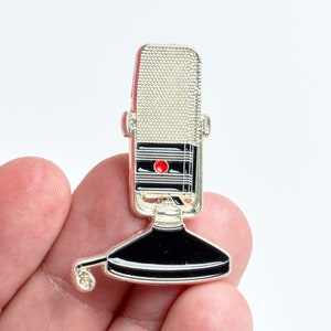 May include: Silver and black enamel pin of a vintage microphone with a red on button.
