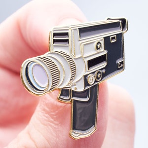 Puede incluir: Un pin de esmalte dorado de una cámara de video vintage. La cámara es de color blanco y negro con una lente dorada.