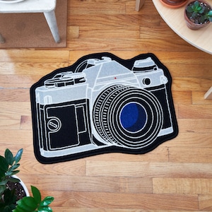 Op de afbeelding: Een zwart-wit tapijt met een vintage camera ontwerp. De camera is wit omrand en heeft een blauwe lens.