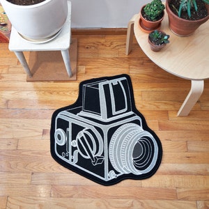 Könnte beinhalten: Ein schwarz-weißer Teppich in Form einer Vintage-Kamera mit detaillierten Konturen. Der Teppich liegt auf einem Holzboden, im Hintergrund sind Topfpflanzen und ein kleiner Tisch zu sehen. Das Kameradesign umfasst ein Objektiv, Einstellräder und einen Sucher.