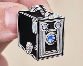 Puede incluir: Un pin de esmalte plateado y negro de una cámara vintage con la palabra "BROWNIE" impresa en la parte delantera.
