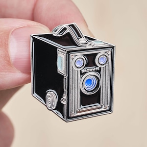 Puede incluir: Un pin de esmalte plateado y negro de una cámara vintage con la palabra "BROWNIE" impresa en la parte delantera.