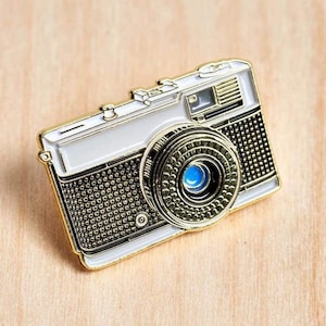 Op de afbeelding: Een gedetailleerde emaille pin in de vorm van een vintage camera. De pin heeft een witte behuizing met gouden randen en een zwarte textuur voorkant. De cameralens heeft een blauw centrum. De pin is geplaatst tegen een lichte houtnerf achtergrond.