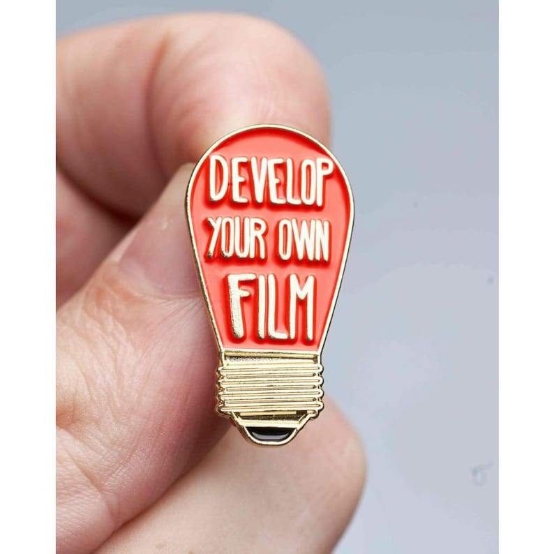 Może przedstawiać: Emaliowana przypinka w kształcie czerwonej żar&oacute;wki z pozłacanym obramowaniem. Przypinka zawiera napis "Develop Your Own Film".