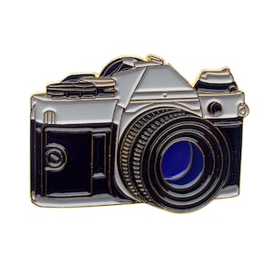 Könnte beinhalten: Ein detaillierter Emaille-Pin in Form einer Vintage-Kamera. Der Pin hat ein schwarz-weiß-silbernes Farbschema mit goldenen Akzenten. Das Objektiv hat eine blaue Mitte. Ein tolles Accessoire für Fotografen.