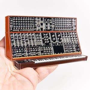 Könnte beinhalten: Eine braun-schwarze Emaille-Anstecknadel in Form eines modularen Synthesizers mit Tastatur. Das Design zeigt ein detailliertes schwarzes Bedienfeld mit zahlreichen Reglern, Schaltern und Patch-Punkten auf einem braunen Rahmen mit passender Basis.