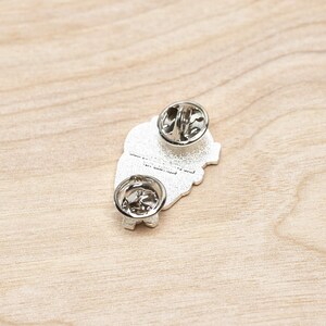 May include: Silver enamel pin featuring a cartoon illustration of a heart with a face and the text "I love you to the moon and back". The pin has a safety clasp on the back.