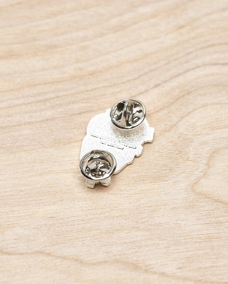 May include: Silver enamel pin featuring a cartoon illustration of a heart with a face and the text "I love you to the moon and back". The pin has a safety clasp on the back.