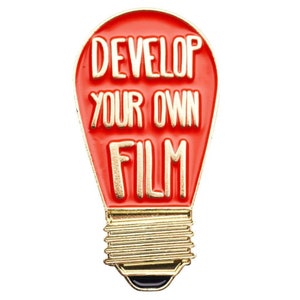 Może przedstawiać: Emaliowana przypinka w kształcie czerwonej żar&oacute;wki z złotym napisem "Develop Your Own Film".