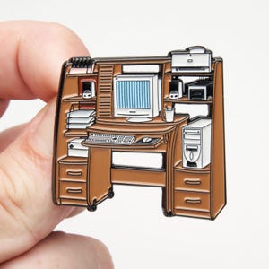 May include: A brown enamel pin shaped like a computer workstation. The desk features a white monitor with a blue screen, a keyboard, a mouse, a tower computer, a printer, speakers, and stacks of paper. The design includes black metal outlines.