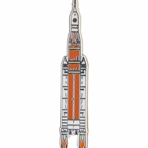 May include: A vertical enamel pin shaped like a space rocket. The design features a central orange cylindrical body flanked by white side panels with black outlines. The top section is white with a small circular window, and the base shows two orange flames.