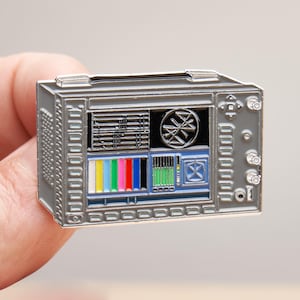 May include: A gray metal enamel pin shaped like a vintage television monitor. The screen displays a grid of test patterns, including a multi-colored vertical bar sequence, a circular graphic, and various technical data readouts with knobs on the side.