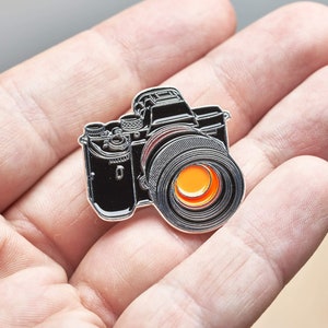 Könnte beinhalten: Eine silberne und schwarze Emaille-Anstecknadel mit einer Kamera und einem orangefarbenen Objektiv. Die Nadel befindet sich in der Handfläche einer Hand.