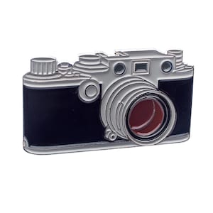 May include: A silver and black camera pin with a red lens. The camera has a detailed design and is a great addition to any collection.