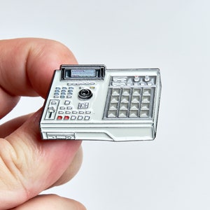 May include: A white and silver music production machine pin. The pin features a detailed design of a vintage sampler with buttons, knobs, and a display screen. The pin is held between two fingers.
