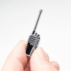 Puede incluir: Un pin de esmalte metálico en tono plateado con forma de radio bidireccional portátil. El diseño presenta una antena larga, una rejilla de altavoz con líneas horizontales y dos perillas pequeñas en la parte superior, delineado en metal plateado pulido.