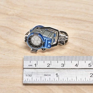May include: A silver and blue enamel pin of a movie studio spotlight. The pin is about 1 inch wide and is sitting on a wooden surface next to a ruler.